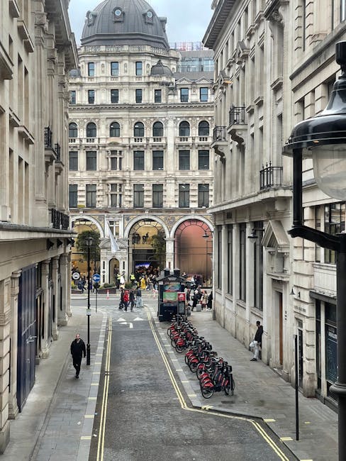 A narrow city street lined with tall, classical-style buildings featuring ornate stone facades and decorative balconies. The street has a smooth asphalt surface with a double yellow line along its edg