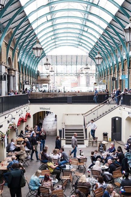 The image depicts the interior of a bustling marketplace with a high, curved glass ceiling supported by green-painted metal arches, allowing natural light to flood the space. Inside, there are multipl