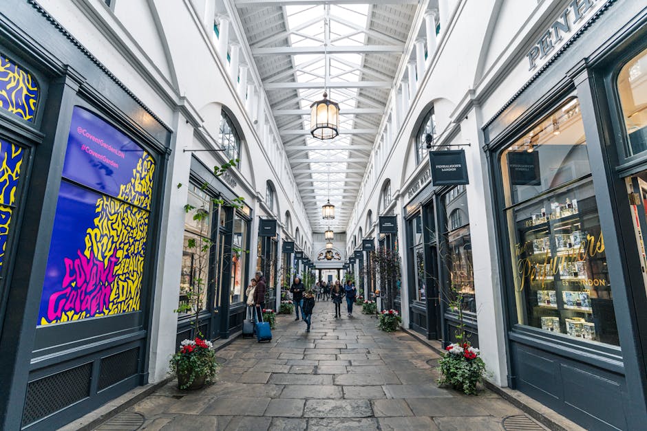 The image depicts the interior of a bustling marketplace with a high, curved glass ceiling supported by green-painted metal arches, allowing natural light to flood the space. Inside, there are multipl